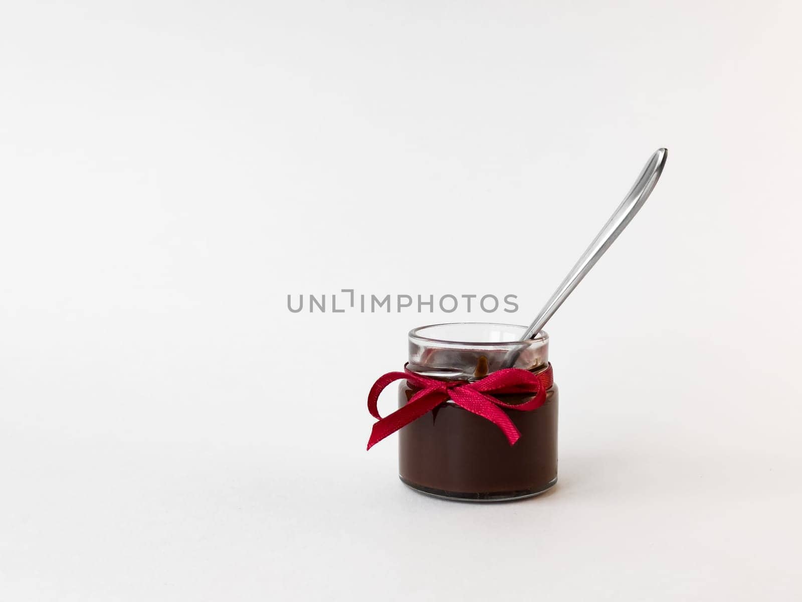 Small glass jar of chocolate spread with red ribbon and spoon on white background.