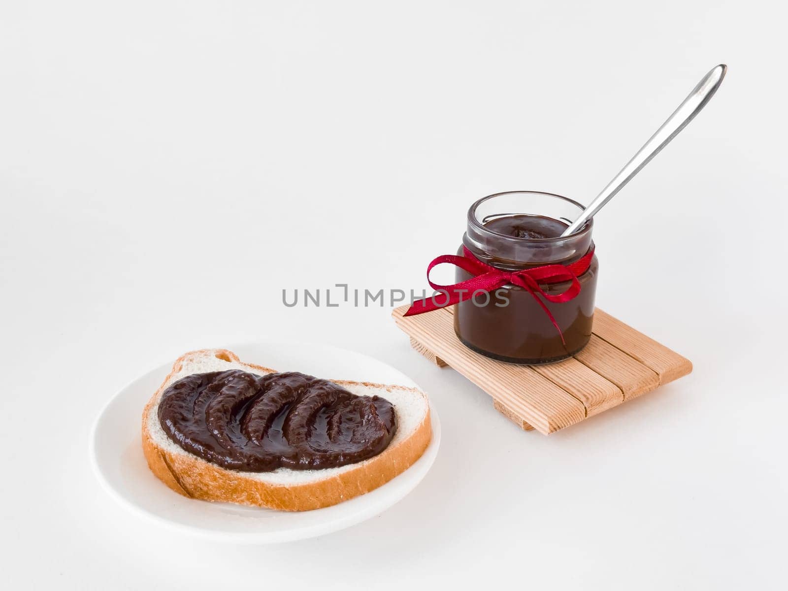 Delicious chocolate spread on bread with jar and red ribbon on white background.