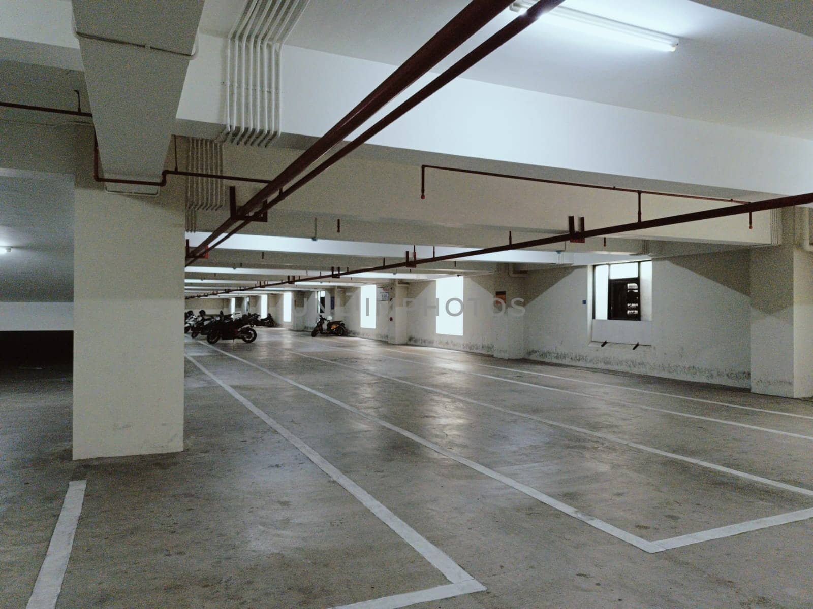 Empty underground parking garage featuring concrete flooring and bright overhead lighting. Spacious area with marked parking lines, suitable for vehicles. Industrial setting with structural beams in view.