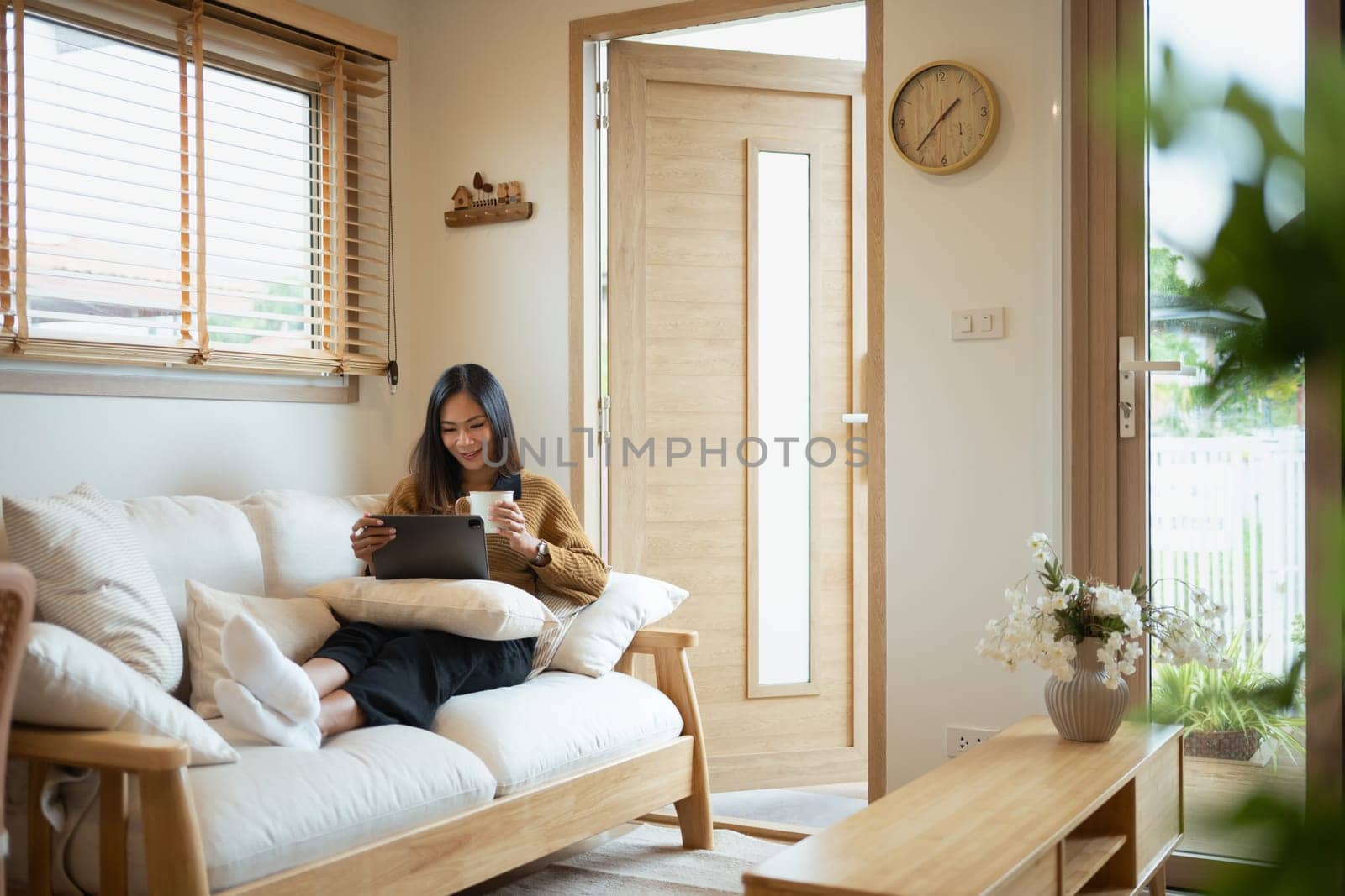 Relaxed young woman drinking hot tea and using digital tablet in living room.
