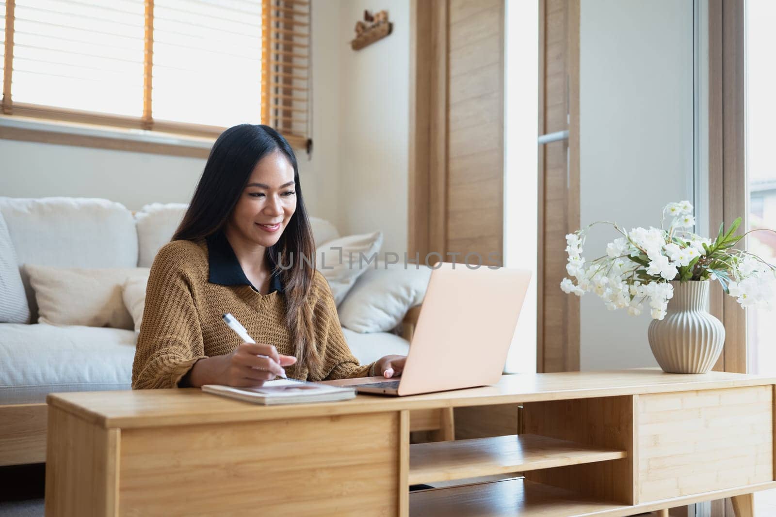 Charming young woman working remotely with laptop and writing notes.