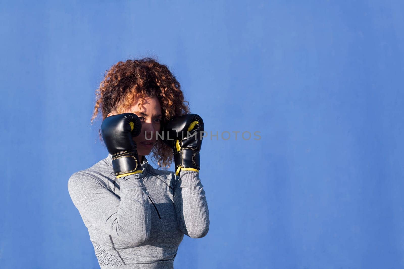 determined adult female boxer with curly hair ready to fight on boxing gloves during workout on blue background, concept of strength and healthy lifestyle, copyspace for text