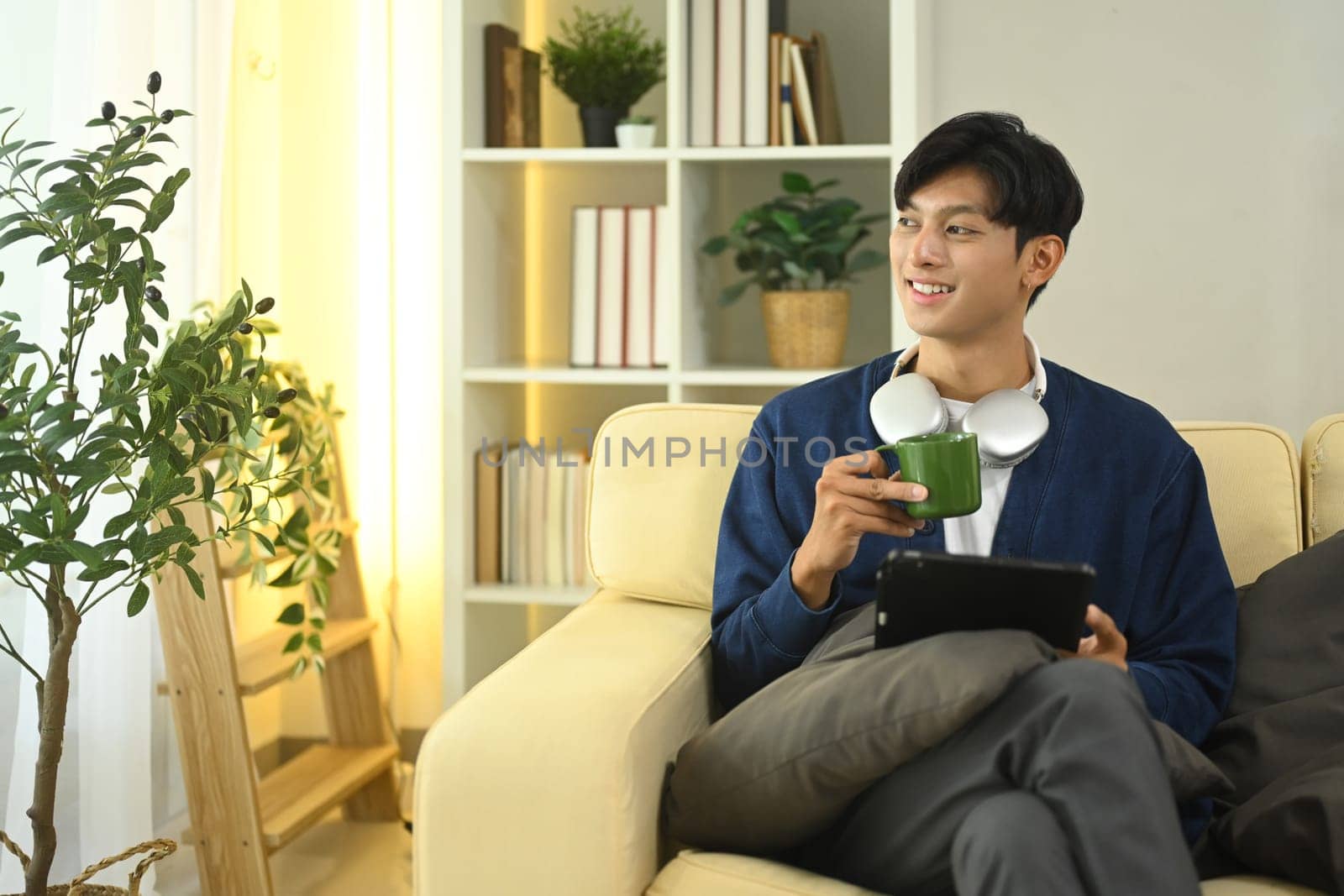 Positive young asian man sipping coffee and using digital tablet on sofa.