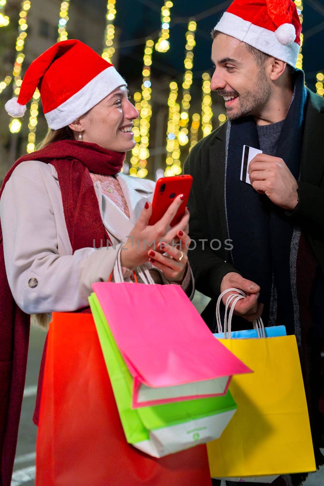 Smiling couple wearing Santa hats, standing under festive Christmas lights. They look at each other warmly, creating a joyful holiday atmosphere. One holds a phone, capturing memories.
