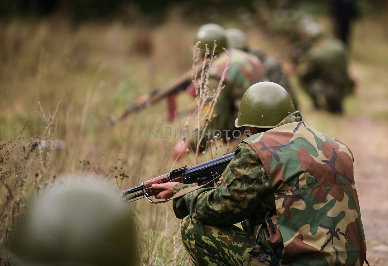 Soldiers in camouflage gear are positioned in a grassy area, focused on training exercises involving shooting while wearing helmets and engaging in tactical maneuvers.
