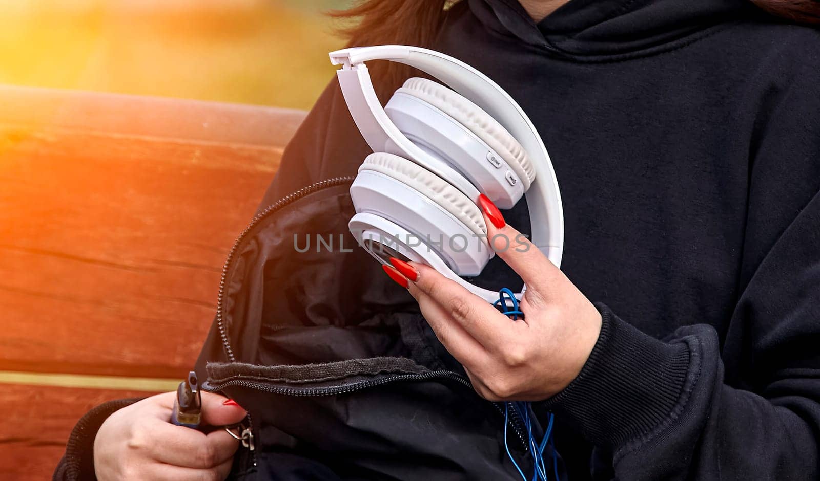 A woman wearing a black hoodie sitting on a bench in a sunny park, holding white over-the-ear headphones with a cord. The background is blurry, creating a serene outdoor setting.