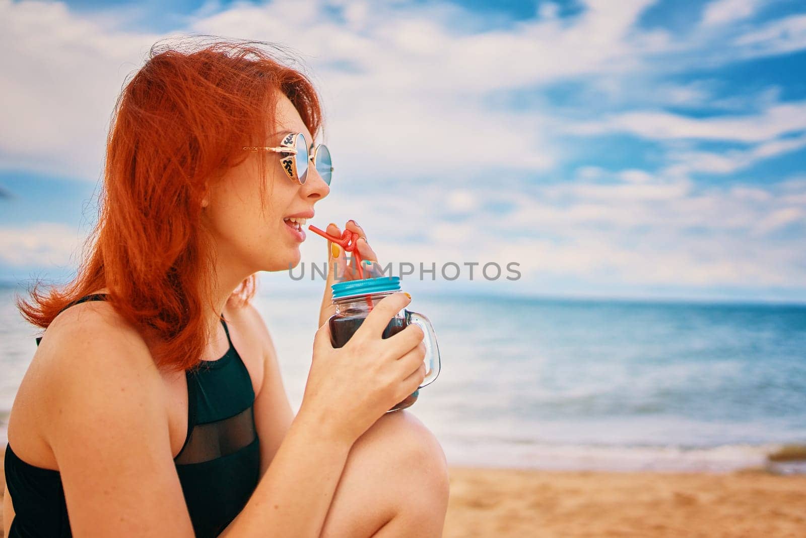 Calm lady in a swimsuit is sitting on beach with a cocktail and look away. Tropical alcohol beverage. Summer day with cloudy sky. Red-haired woman with sunglasses on vacation. Holidays at the sea.