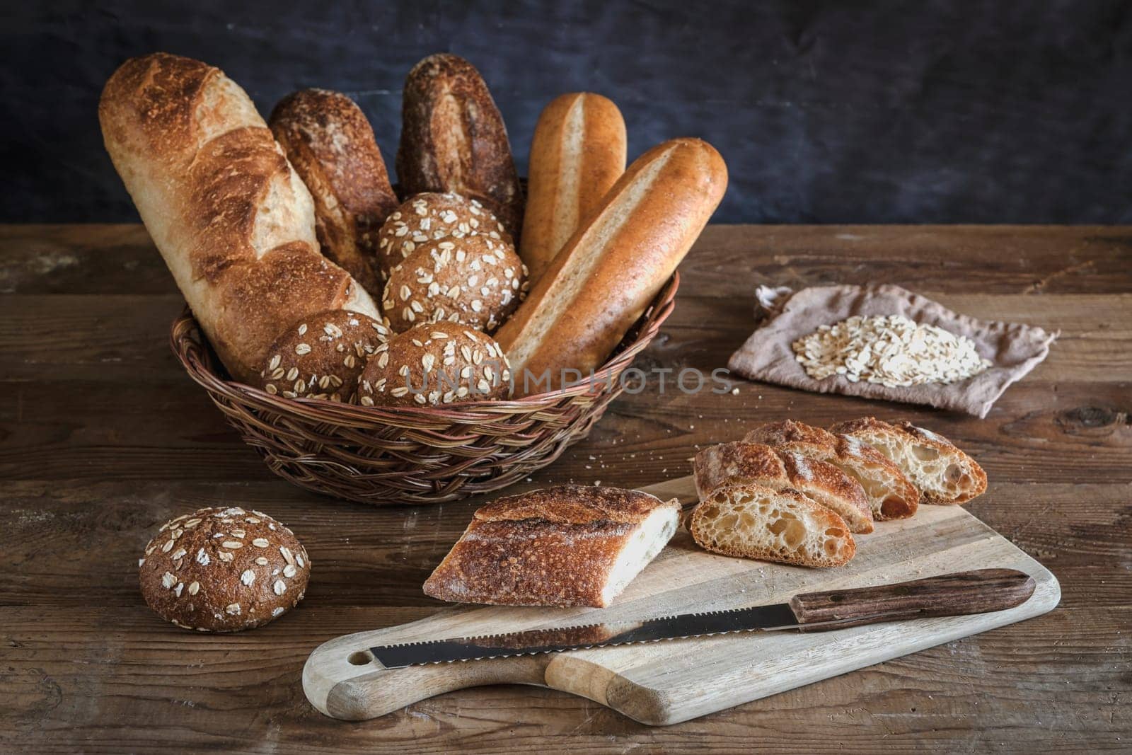 An assortment of bread in a basket and a baguette on the cutting board.