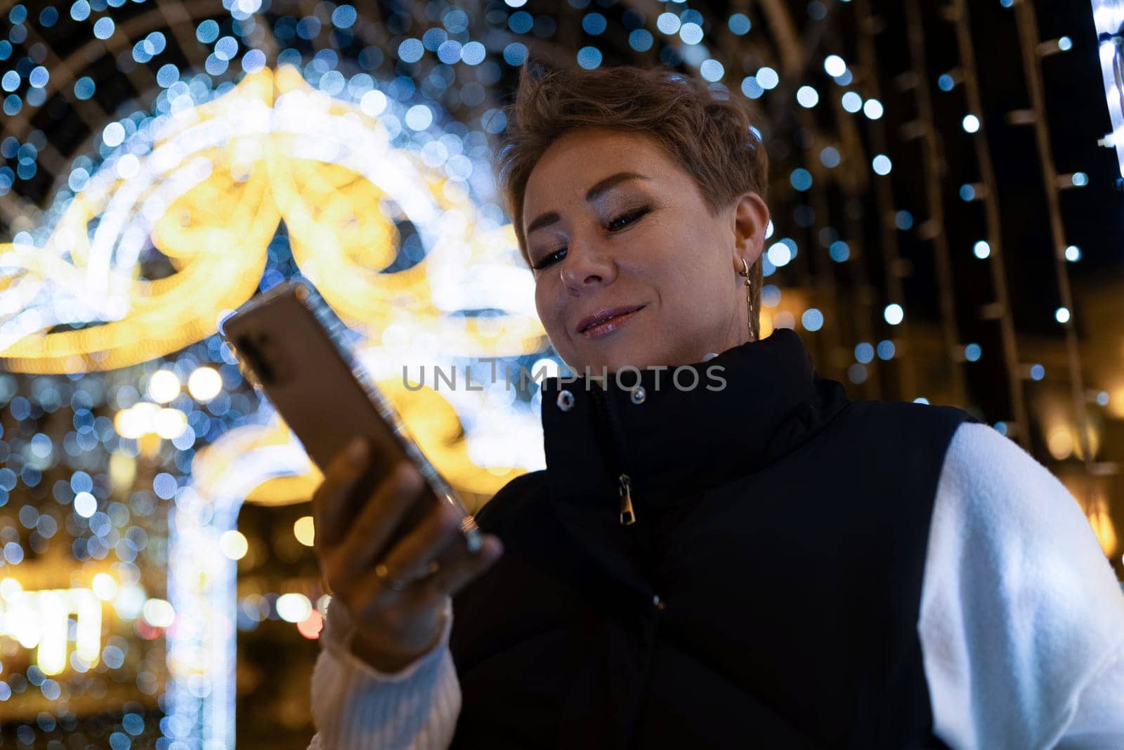 A woman with short brown hair walks through a city at night. She is looking at her phone and is smiling. There are string lights in the background, out of focus.