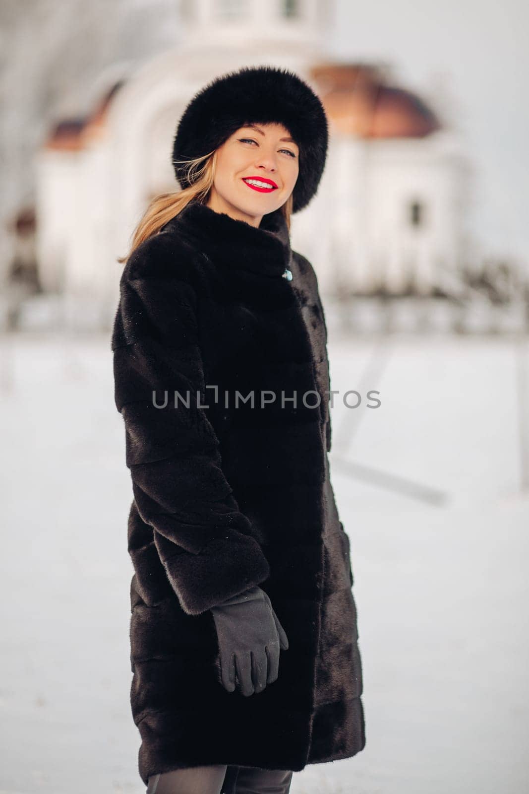 A beautiful woman in a black fur coat and a black hat has a lot of snow in winter, she is wearing red lipstick, smiling.