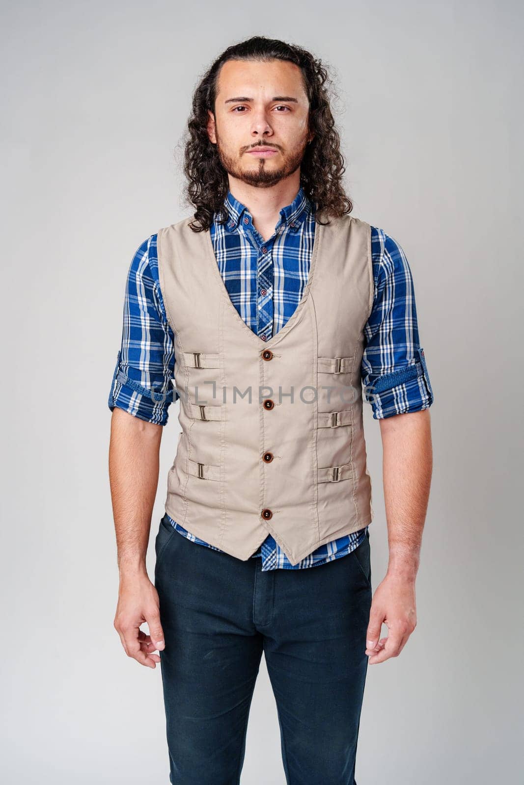 Young man in plaid shirt and vest poses confidently against a neutral background in a studio setting