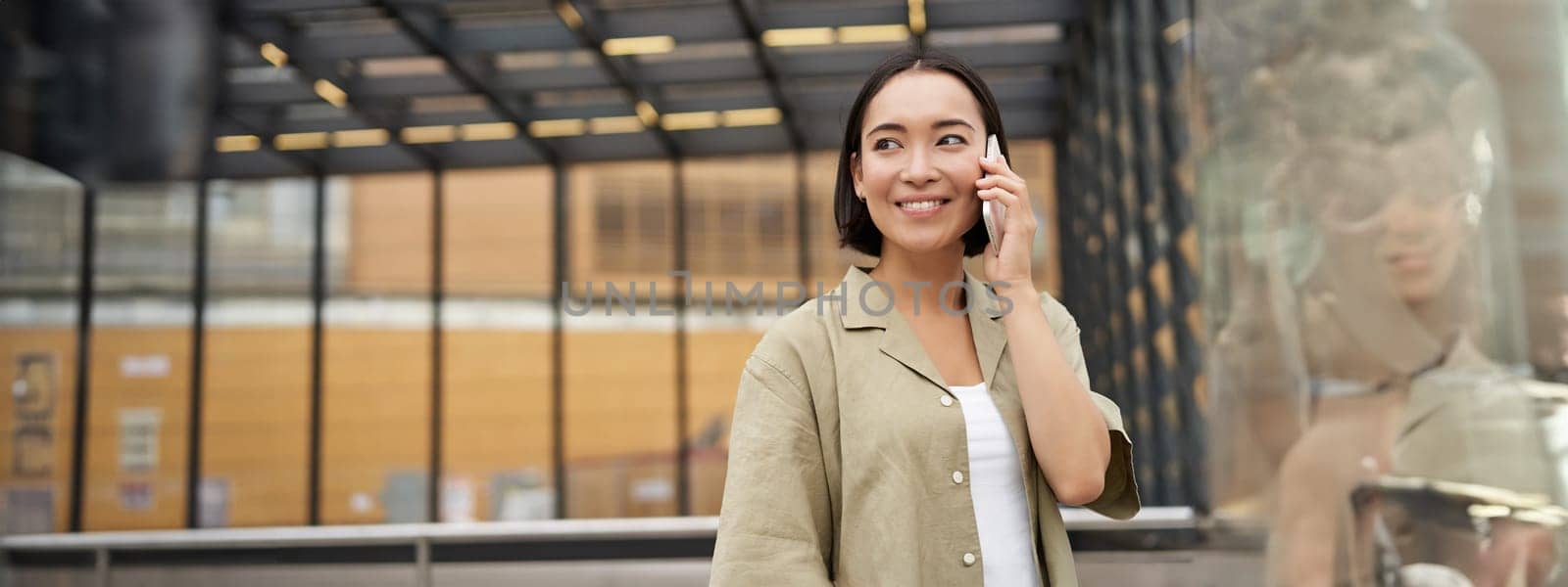 Happy asian woman talking on mobile phone, standing on bus stop in city. Outdoor shot by Benzoix