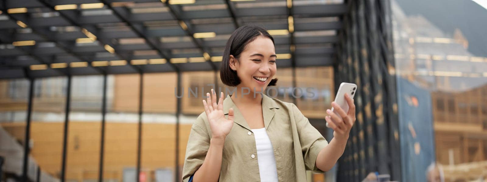 Happy asian girl waves at her smartphone camera, says hello to friend on video chat, calling someone, standing on street by Benzoix