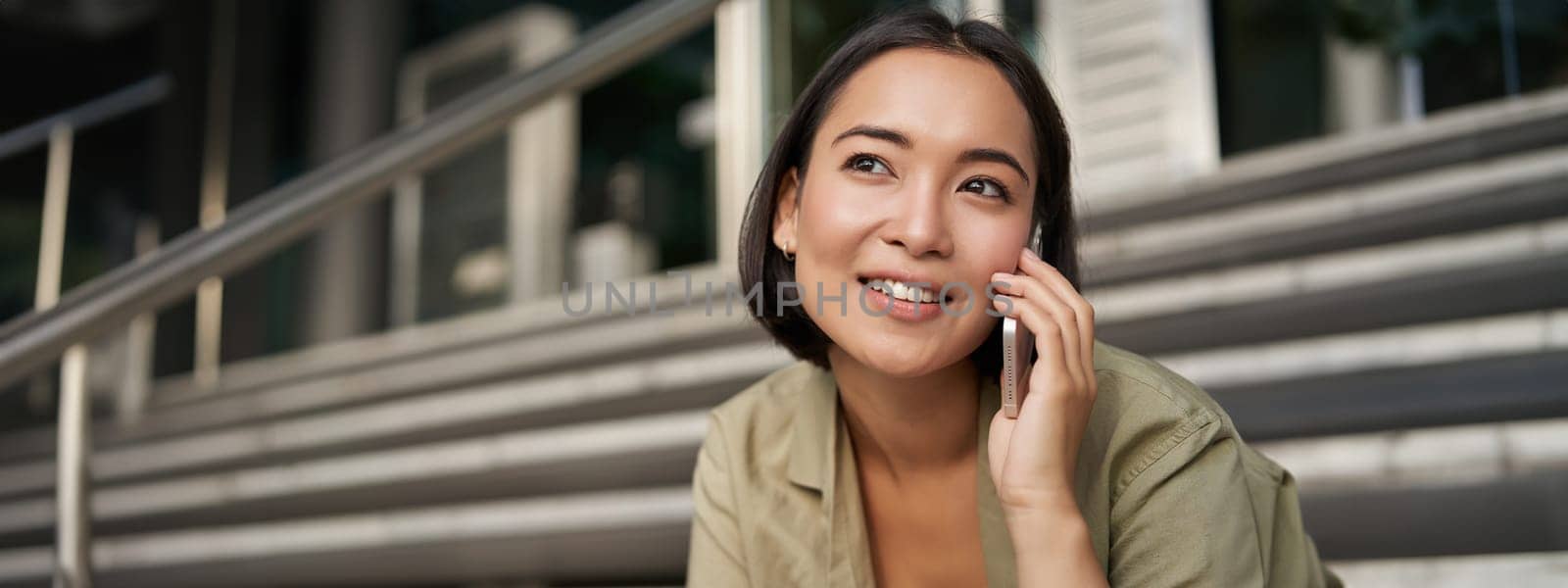 Portrait of asian girl smiles while talks on mobile phone. Young woman calling a friend, sitting on stairs by Benzoix