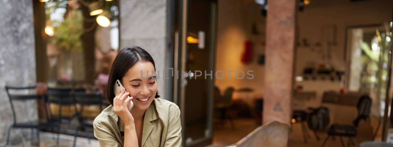 Portrait of young woman talking on mobile phone while typing, using laptop, working remotely from outdoo cafe, doing homework and calling.