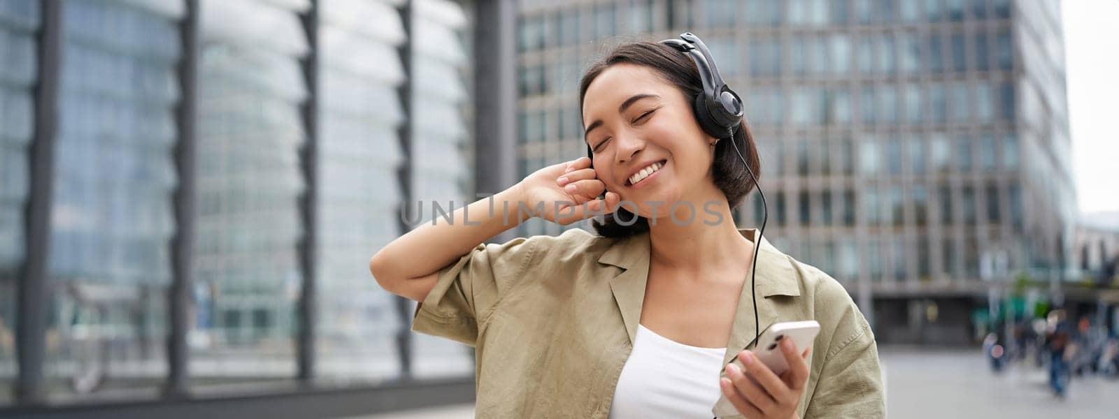 Happy young woman dancing on streets and listening music in headphones, holding smartphone by Benzoix