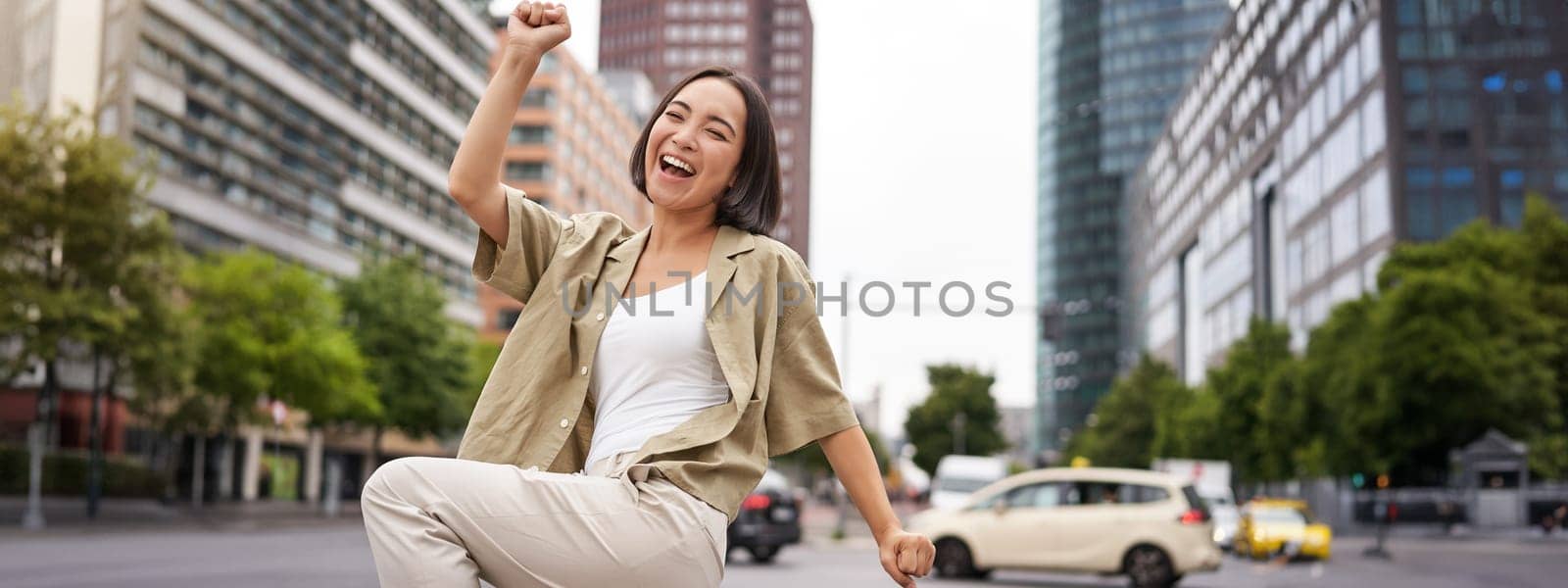 Portrait of happy asian woman, dancing and feeling joy, triumphing, raising hand up in victory gesture, celebrating on streets by Benzoix