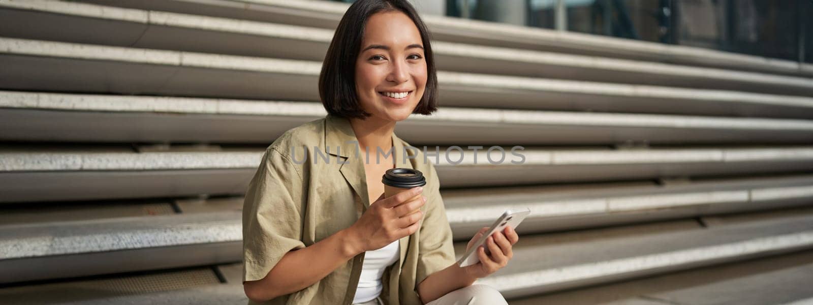 Portrait of asian woman with smartphone, drinks coffee and watches videos on mobile phone. Girl with telephone sits on stairs outdoors by Benzoix