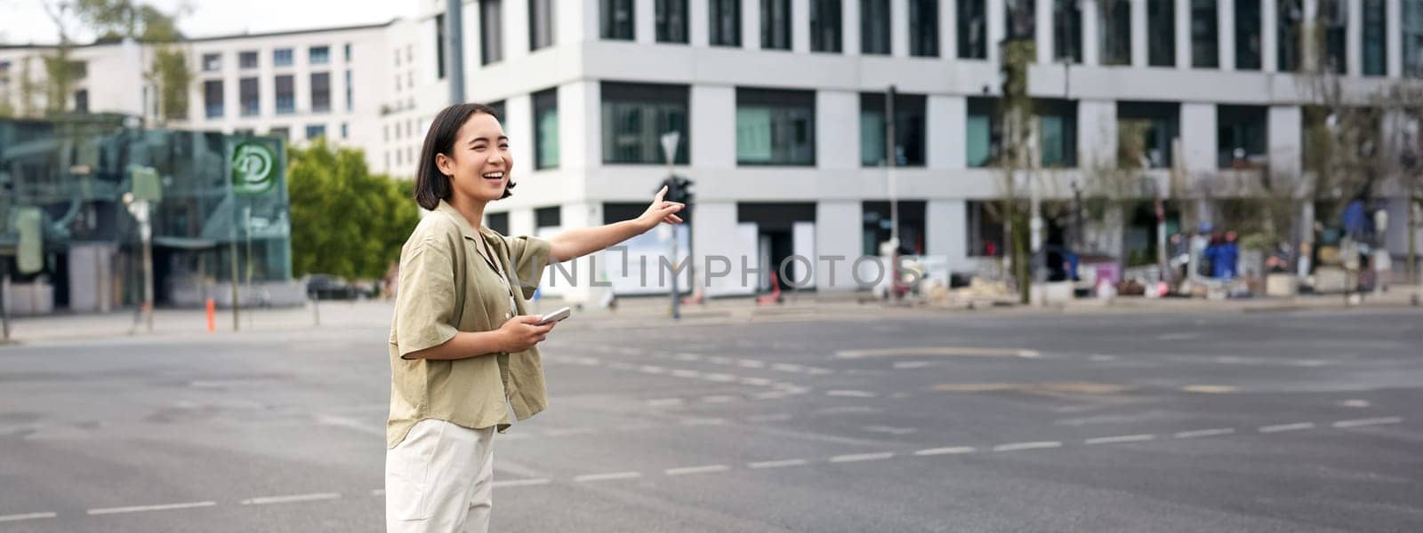 Vertical shot of young woman trying catch taxi, waving at driver on road, holding smartphone with car sharing app, standing on city street by Benzoix