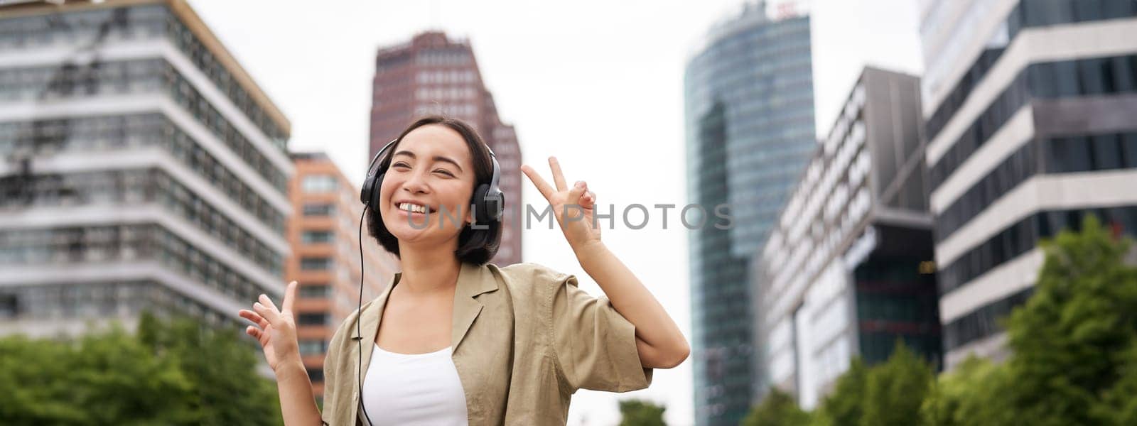 Happy asian woman in headphones, listening music and dancing on street of city centre, smiling with hands up.