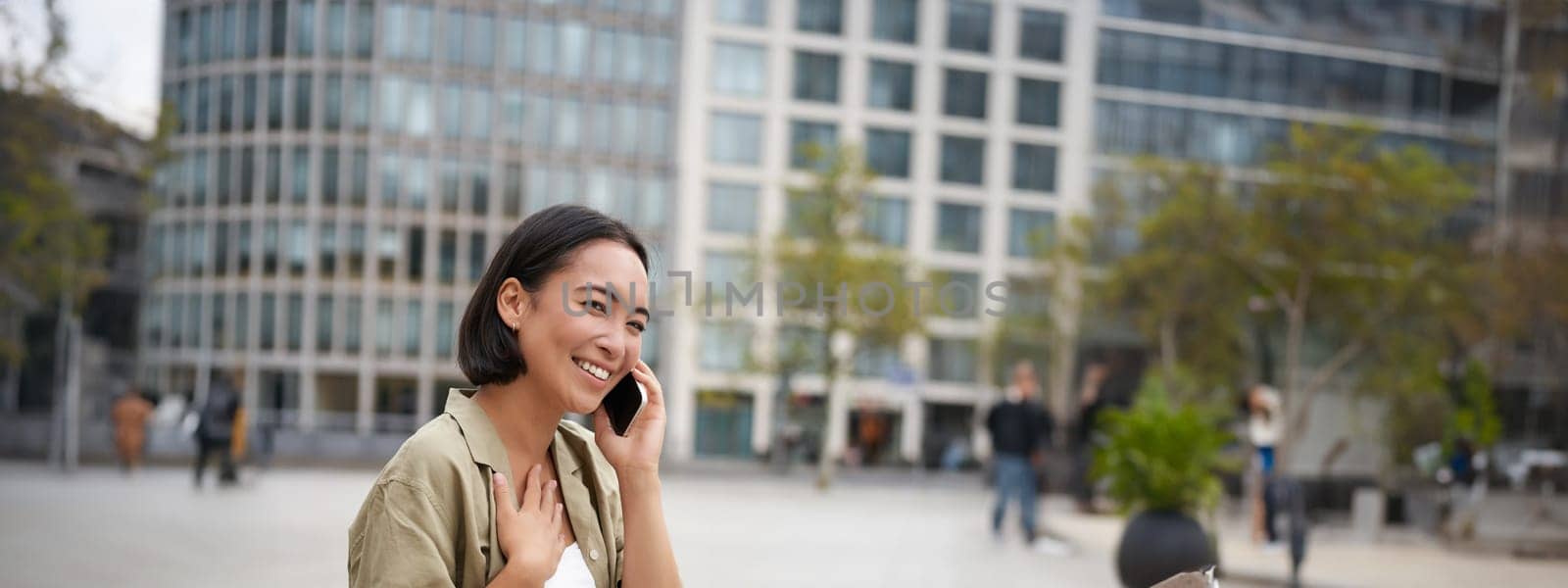 Vertical shot of smiling stylish asian girl, having a telephone conversation and walking on street. Young woman talking on mobile phone.