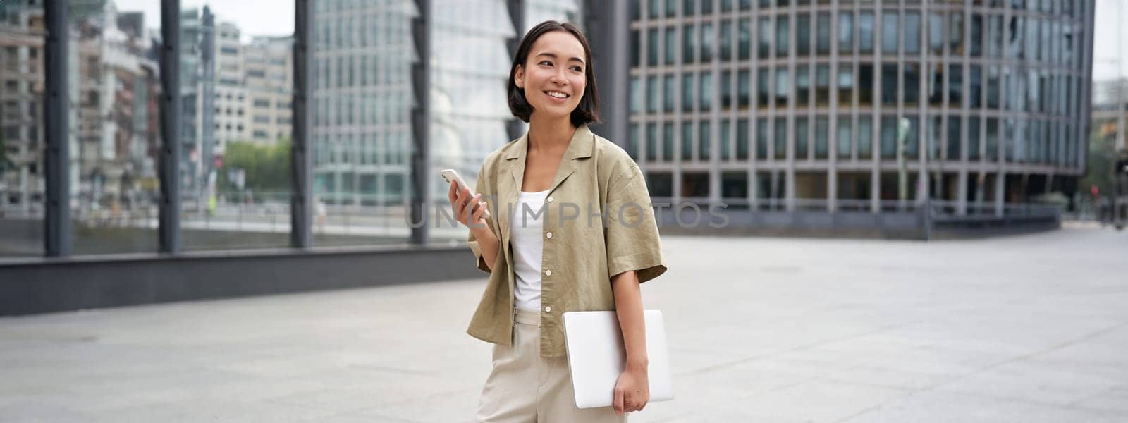 Asian girl with laptop and smartphone, standing on street of city centre, smiling at camera.