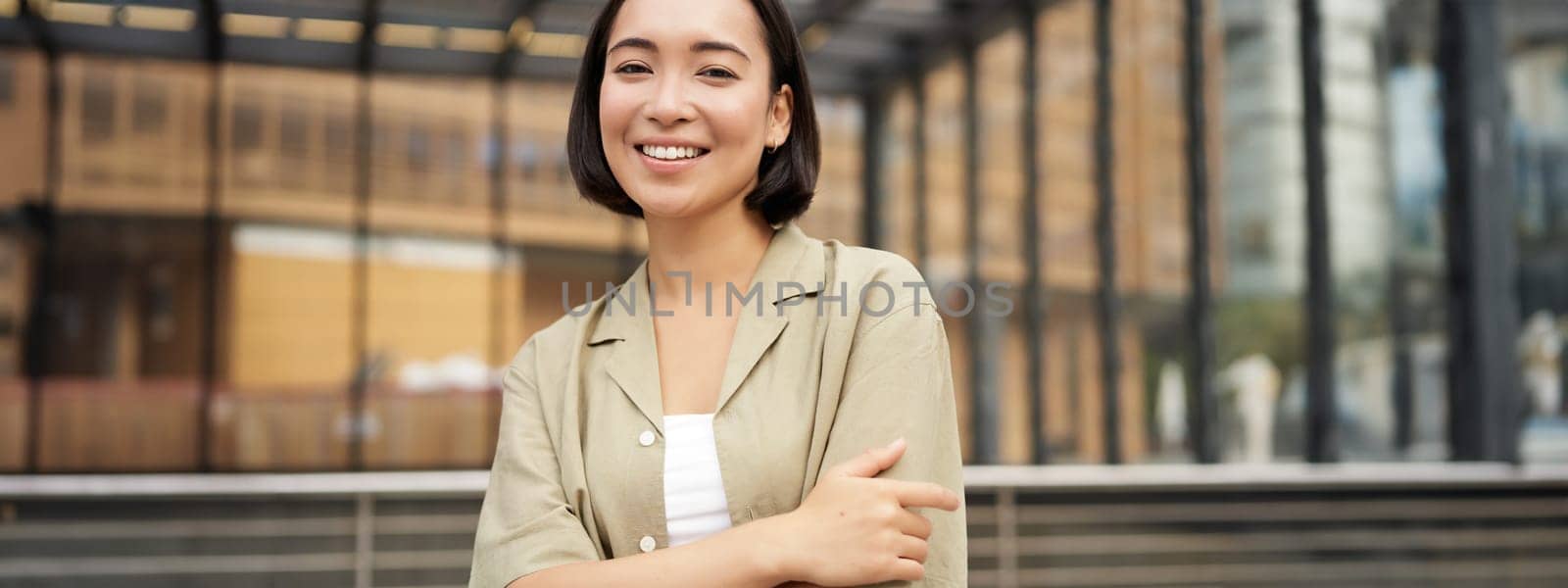 Portrait of young confident female model, girl in casual clothes, posing outside near glass building, smiling at camera.