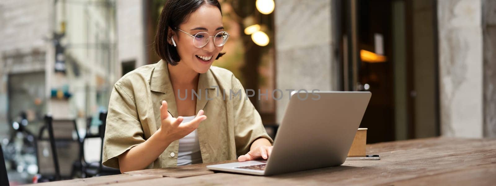 Online meeting. Young urban girl, asian woman talking with laptop, video chat, gesturing, sitting in an outdoor cafe and working remotely by Benzoix