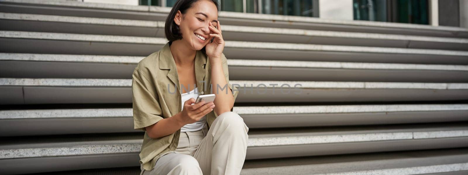Portrait of happy asian girl sits with mobile phone and laughs. Young woman with smartphone being outside by Benzoix