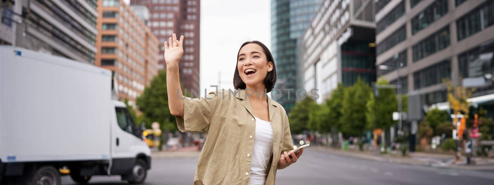 Happy asian girl passing by friend and waving at them on street, saying hello while walking in city, holding smartphone by Benzoix