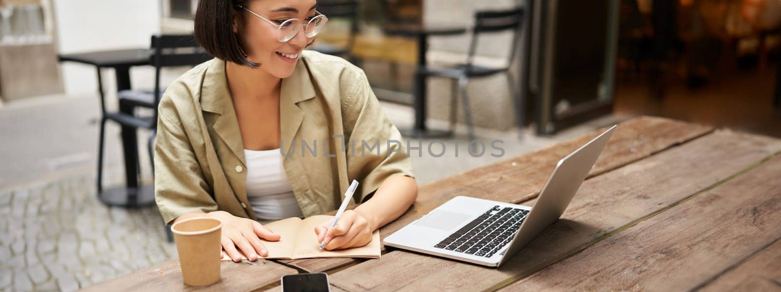 Portrait of concentrated girl doing homework, attending online course, making notes while working on laptop from cafe by Benzoix