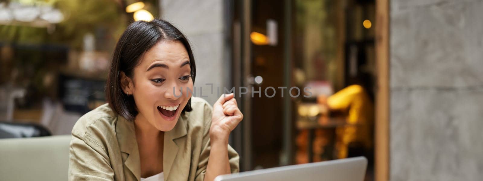 Vertical shot of happy girl talking on video call, looks at laptop, having online meeting, sitting in outdoor cafe by Benzoix