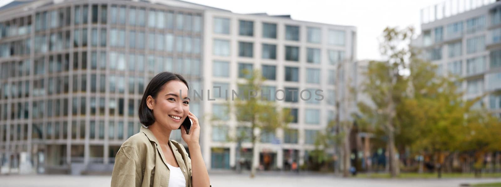 Modern young asian girl talks on mobile phone, uses telephone on city street. Woman smiling while calling someone on smartphone by Benzoix