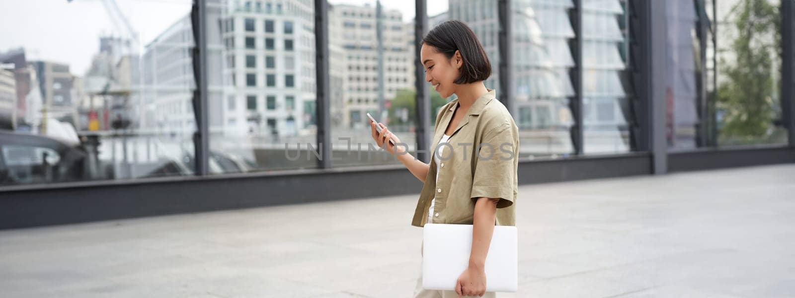 Young asian woman with laptop, walking on street and texting message, smiling while looking at smartphone by Benzoix