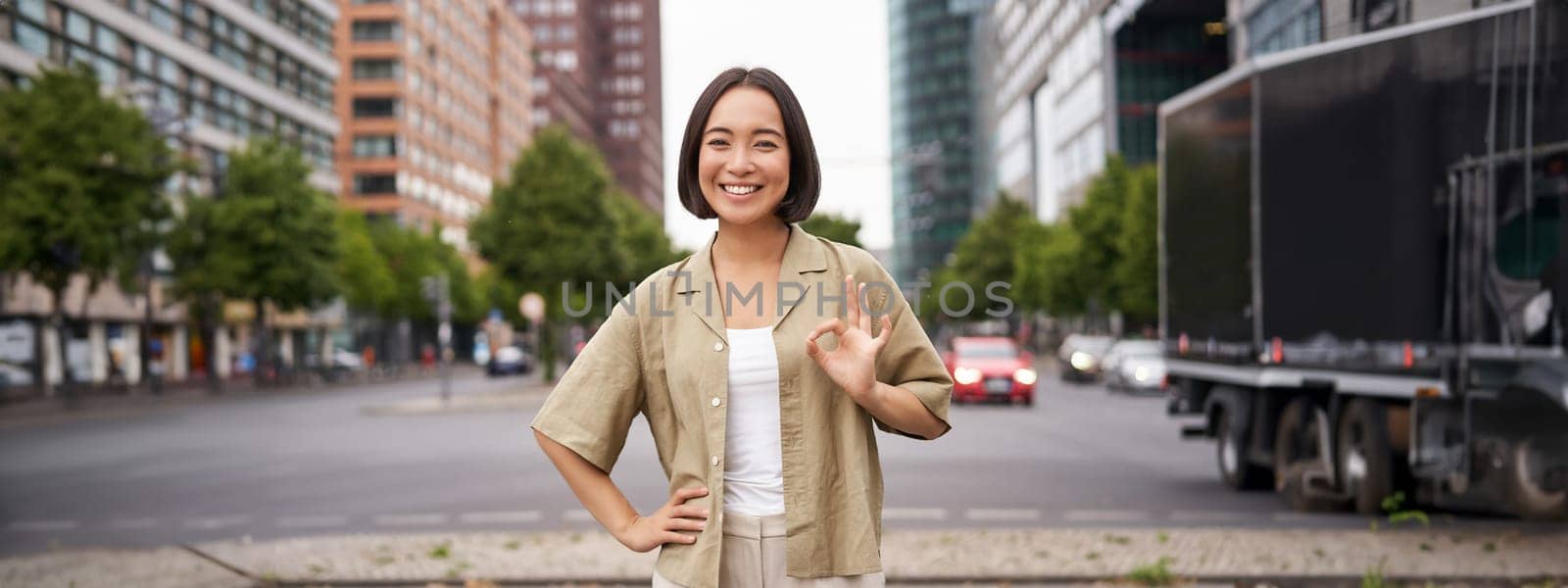 Enthusiastic city girl, shows okay gesture in approval, looking upbeat, say yes, approves and agrees, stands on street.