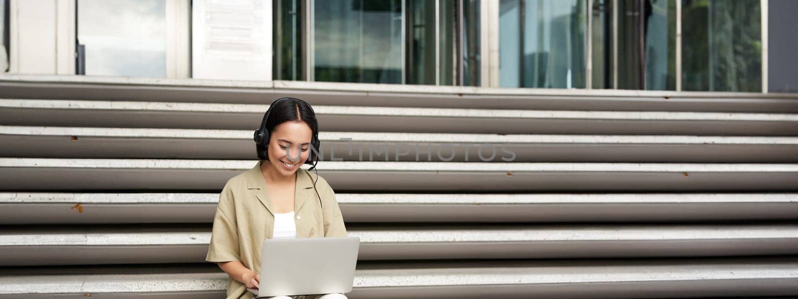 Smiling asian girl in headphones, works on laptop, digital nomad using computer on remote, sitting on stairs outdoors by Benzoix