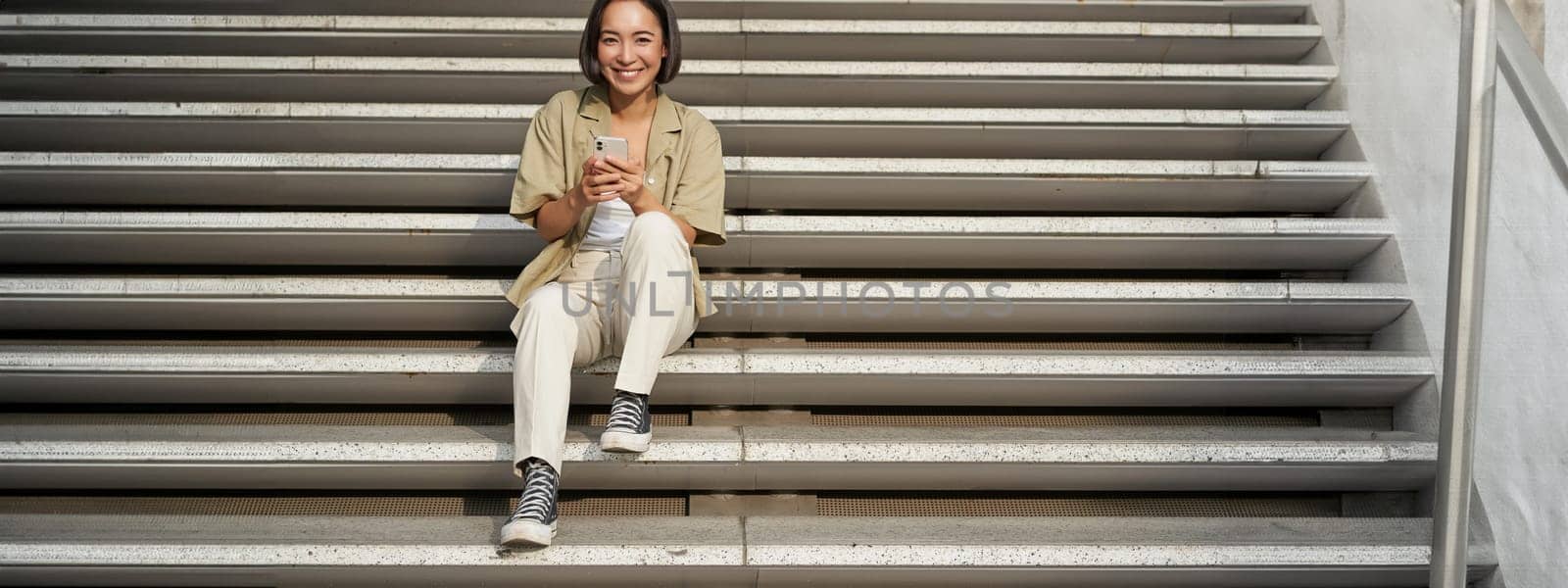 Portrait of smiling asian girl sits on stairs with her smartphone, browsing internet on mobile phone, resting outdoors in city centre by Benzoix