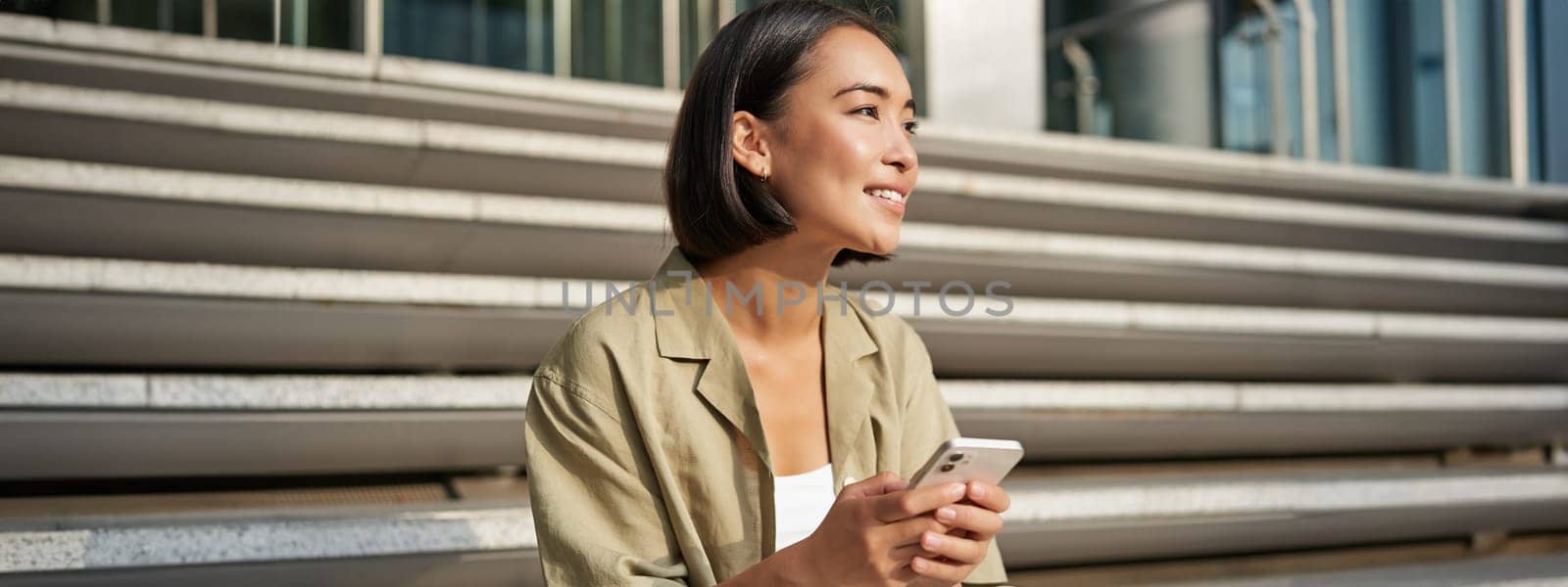 People and technology. Smiling beautiful asian woman sitting on stairs in city, holding mobile phone. Girl with smartphone rests outside by Benzoix