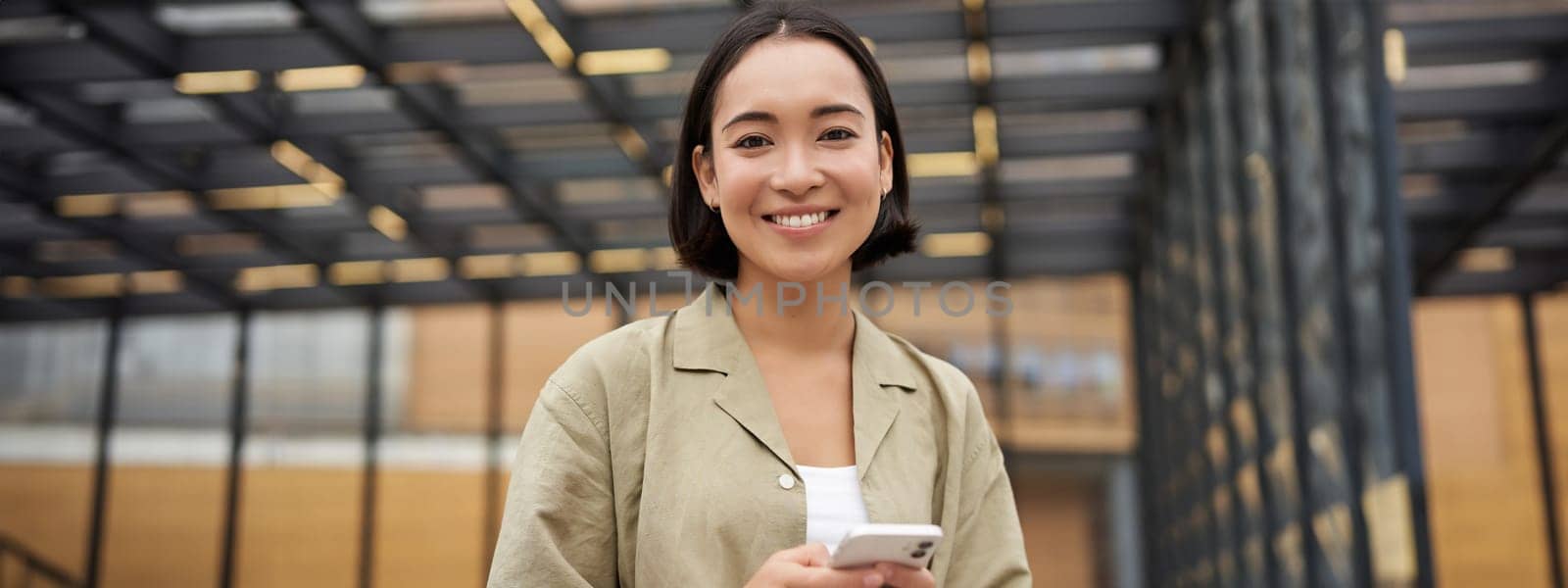 Technology and people. Smiling asian girl with mobile phone, using telephone and walking in city in daylight.