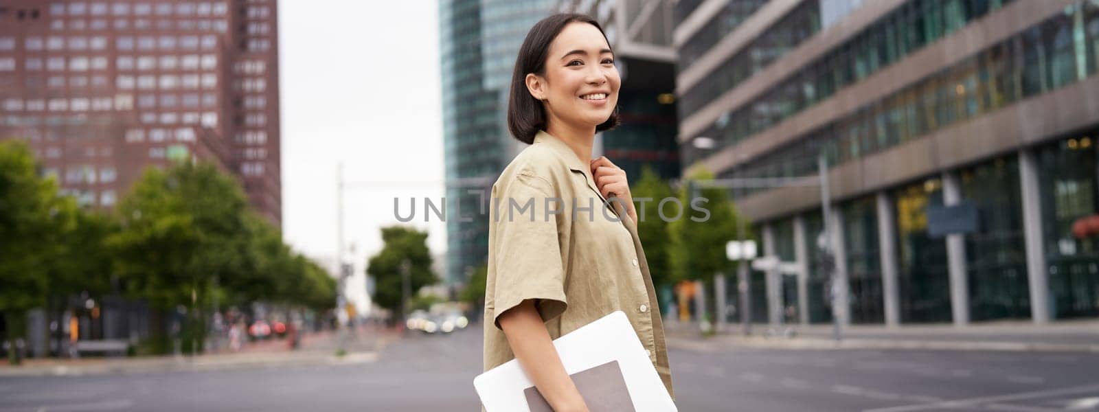 Outdoor shot of asian girl with laptop, going somewhere in city centre, walking on street, going to work.