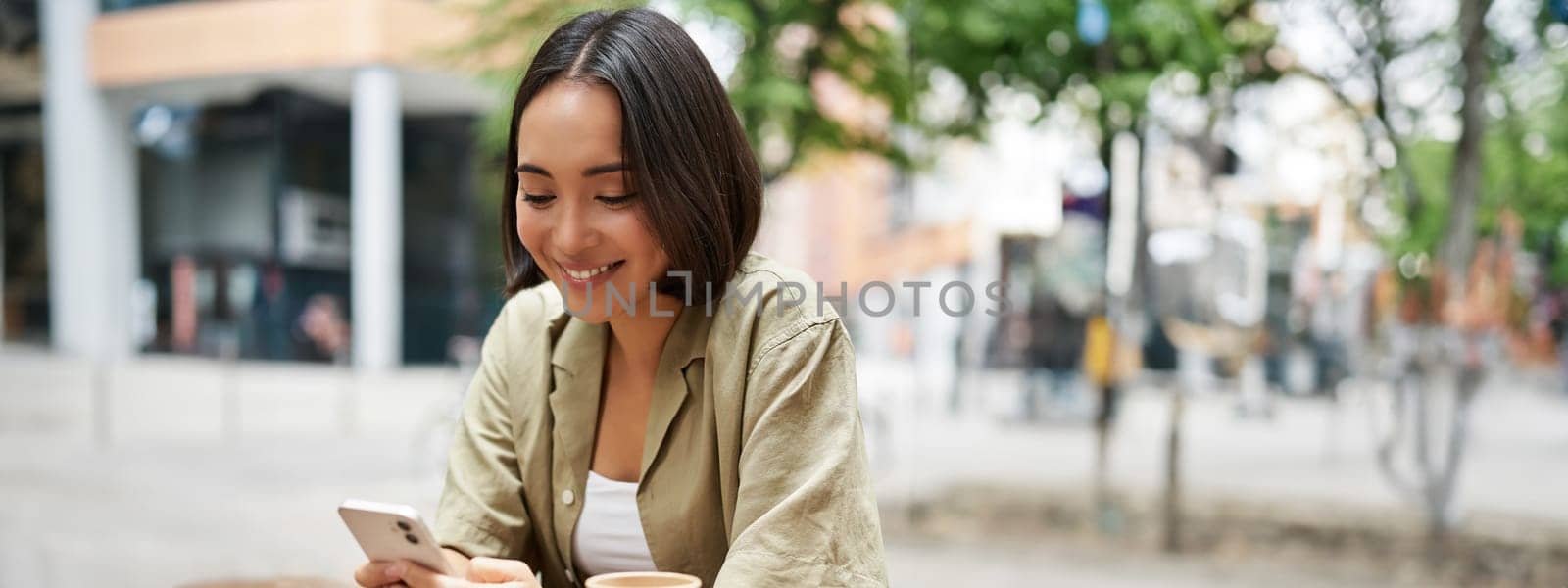 Portrait of young woman enjoying her coffee, drinking takeaway on bench in city, using smartphone by Benzoix