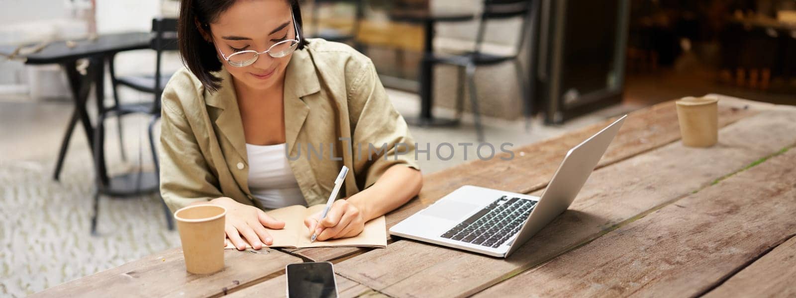 Portrait of concentrated girl doing homework, attending online course, making notes while working on laptop from cafe by Benzoix