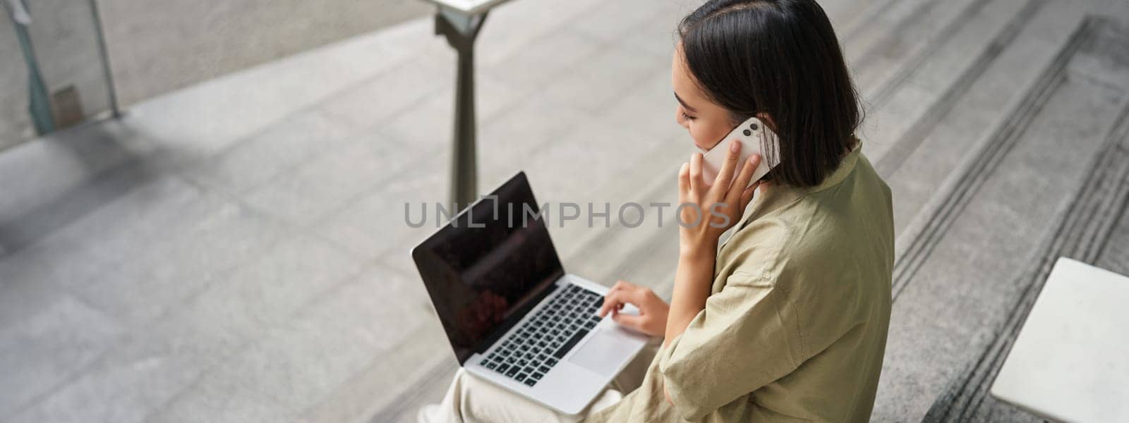 Portrait of asian girl, student sits on stairs with laptop, talks on mobile phone. Young woman makes a telephone call while working on computer.