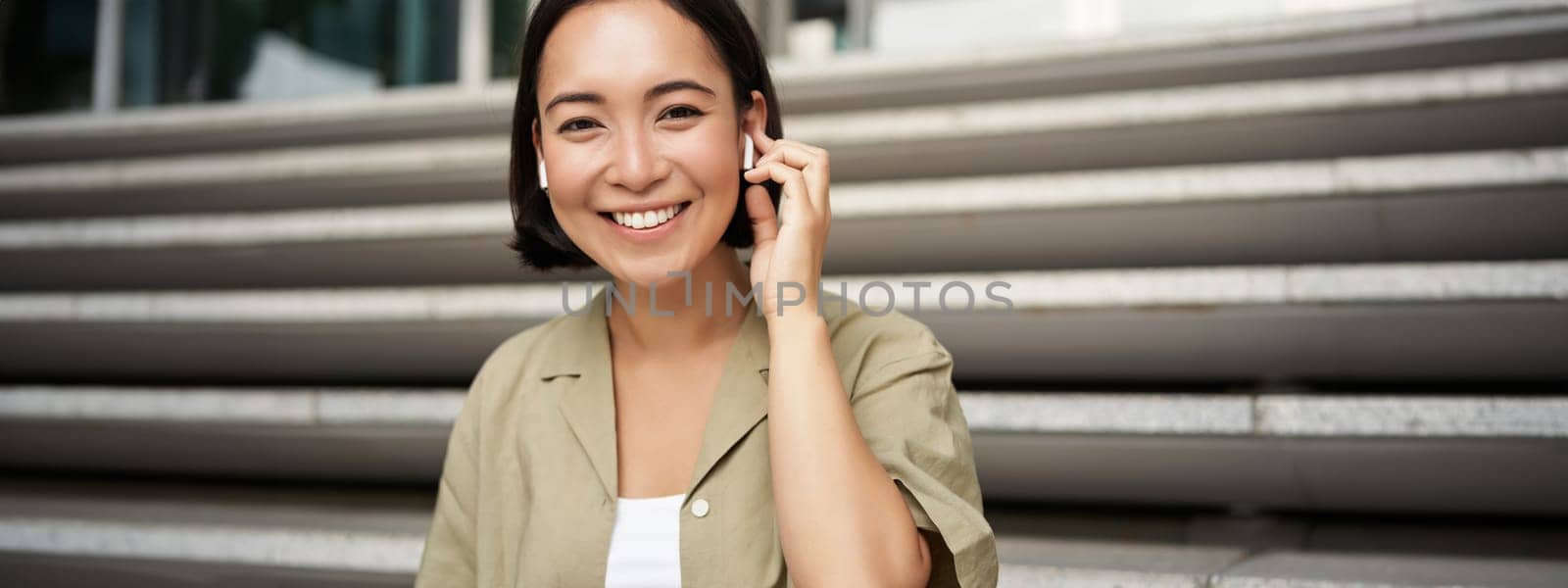Portrait of smiling asian girl listens music, podast in wireless earphones, using headphones outdoors, sitting on street.