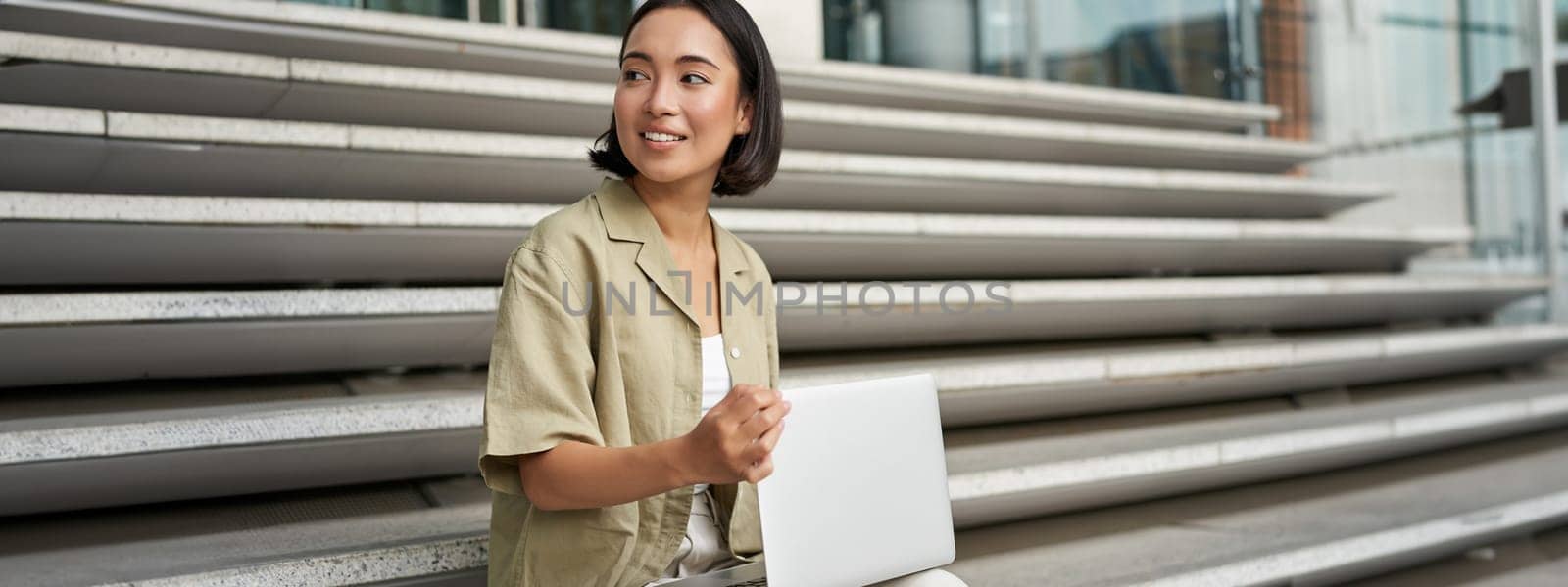 Portrait of young woman student, asian girl using laptop. Asign smiling girl, digital nomad works on her project remotely, sitting on street by Benzoix