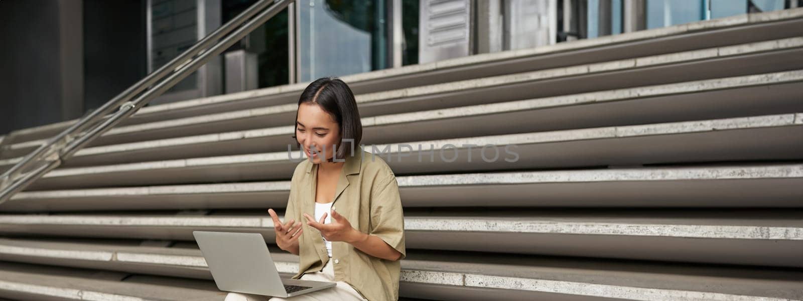 Vertical shot of asian girl student attend online meeting, talking at laptop, video chat, sitting on stairs by Benzoix