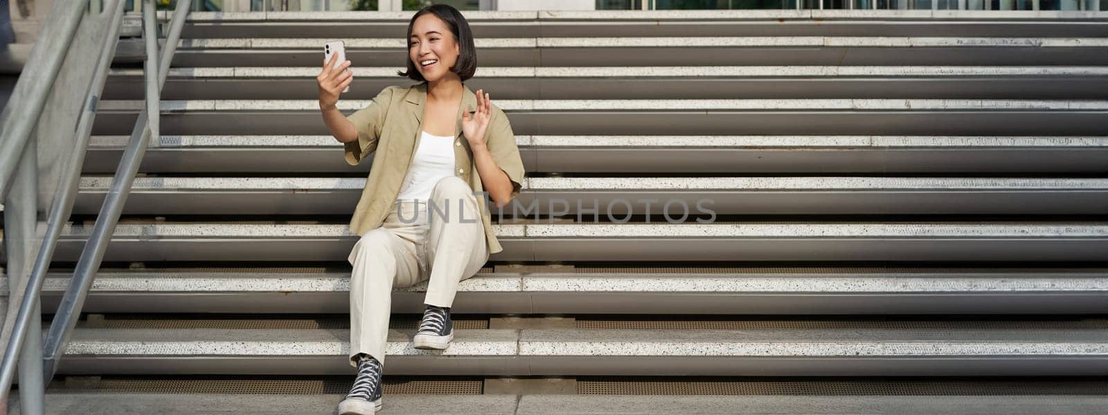 Portrait of asian beautiful girl takes selfie on smartphone. Young korean woman sits on stairs outdoors and makes photos on mobile phone.