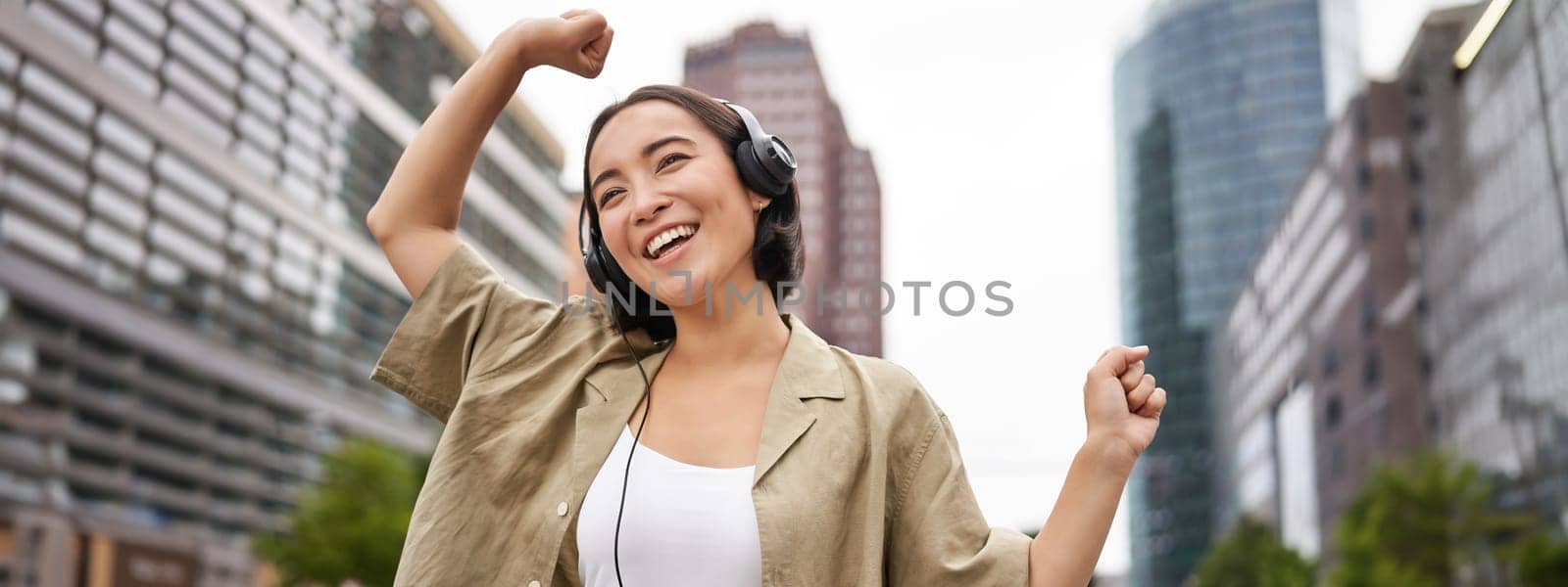 Dancing asian girl listening music in headphones, feeling happy in city, enjoying outdoors.