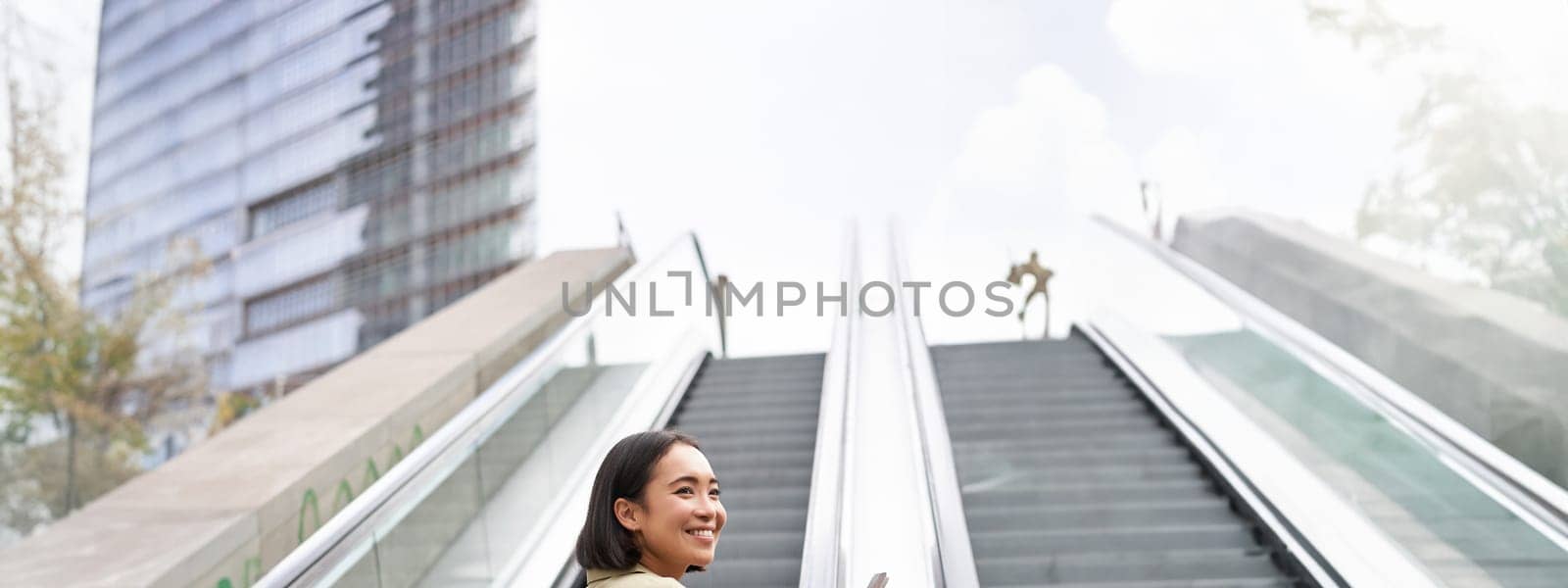 Young asian girl going up on an escalator, holding smartphone, smiling while walking in city. Copy space