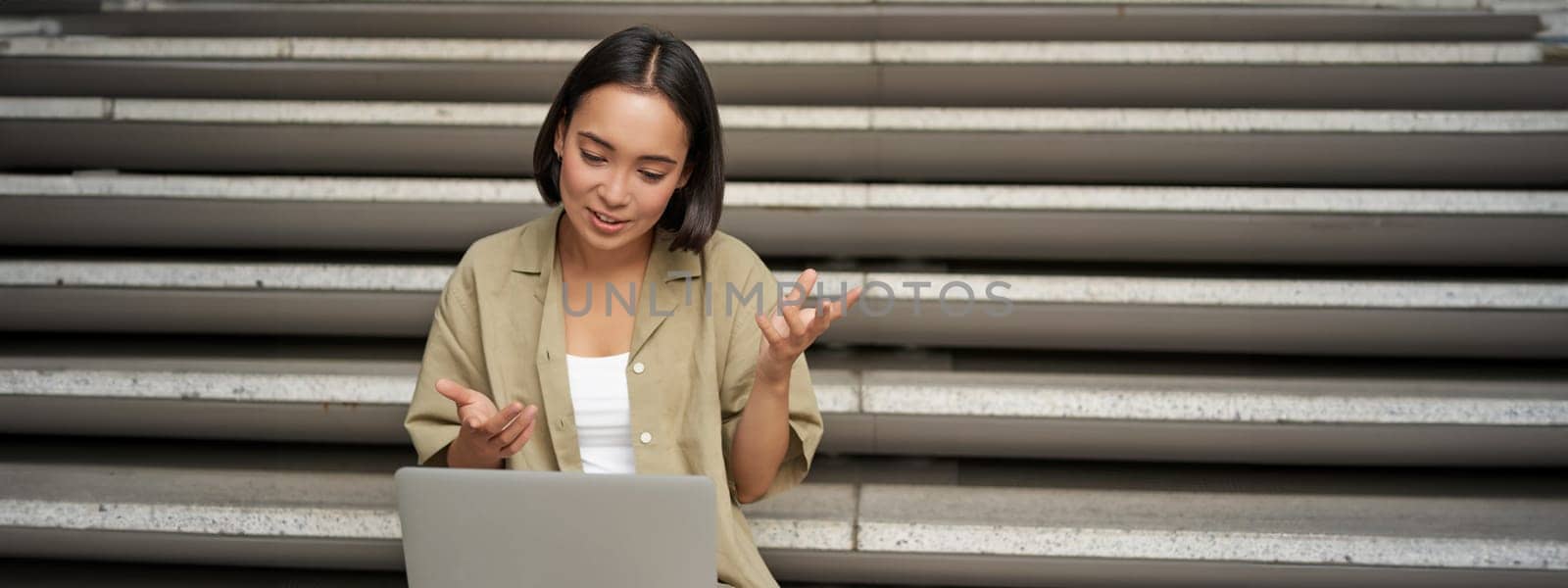 Portrait of young asian girl, student talks at laptop, video chat, speaking during online meeting, sitting outdoors on stairs.